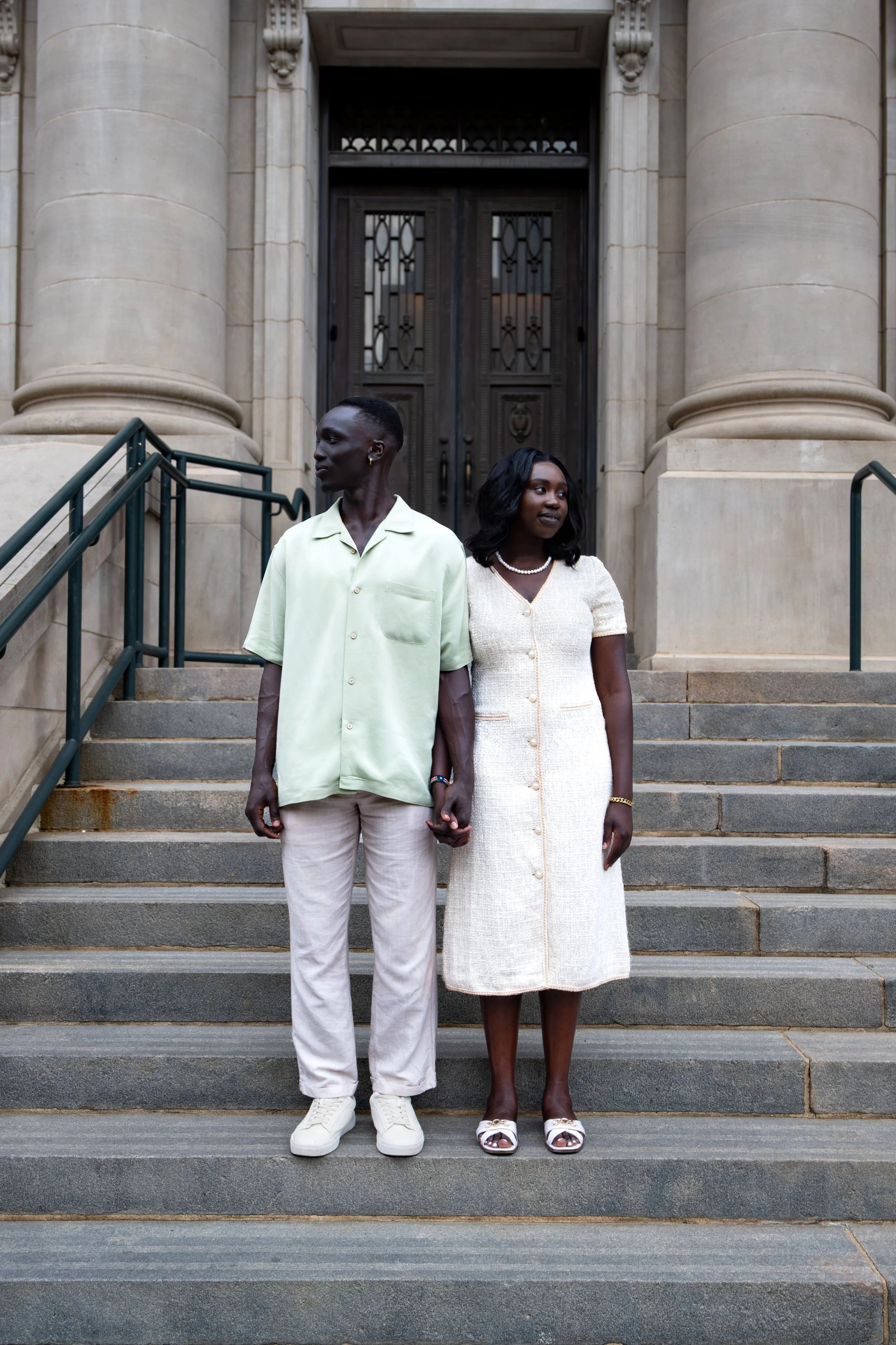 A man and woman holding hands and standing on the steps of a building with large columns and a heavy wooden door in the background.