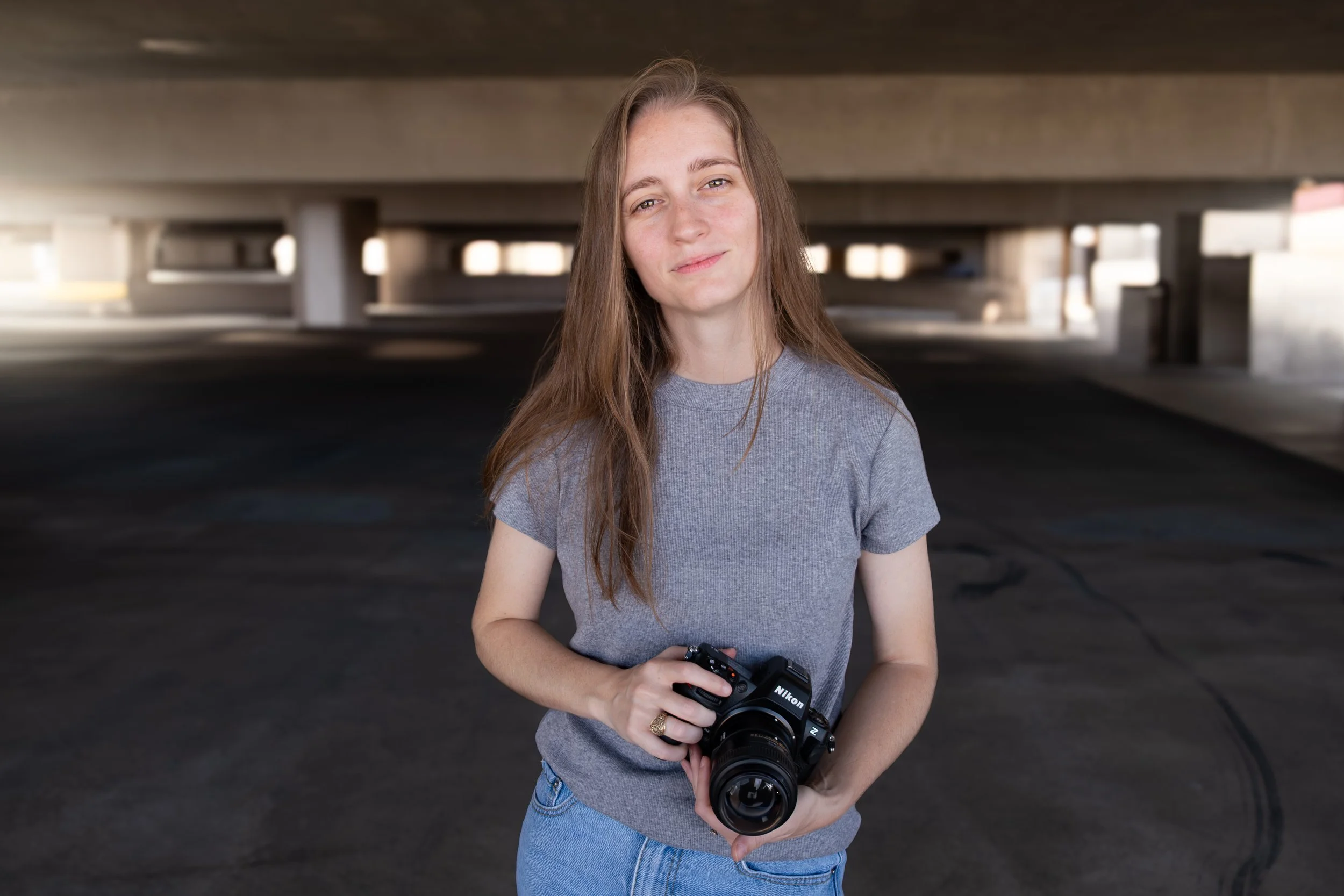 A young woman with long brown hair, wearing a gray t-shirt and blue jeans, is standing under an overpass holding a Nikon camera in her hands. She is smiling slightly and looking at the camera.