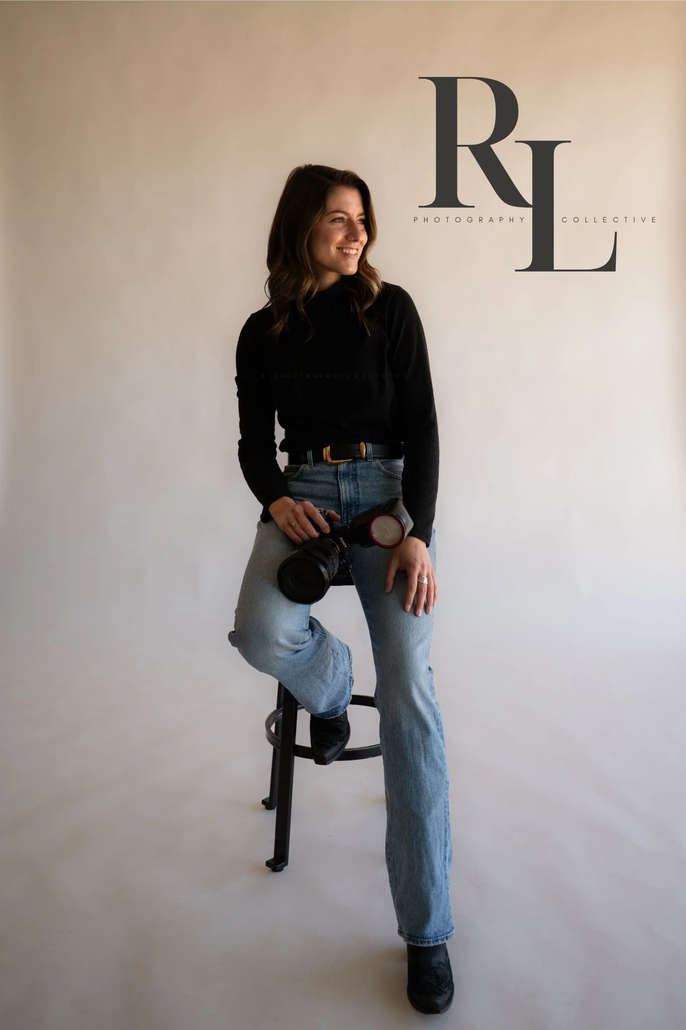 A smiling woman with dark hair, wearing a black turtleneck, blue jeans, and black shoes, sitting on a stool and holding a camera, in front of a plain light-colored background with the logo 'RLC Photography Collective' on the wall.