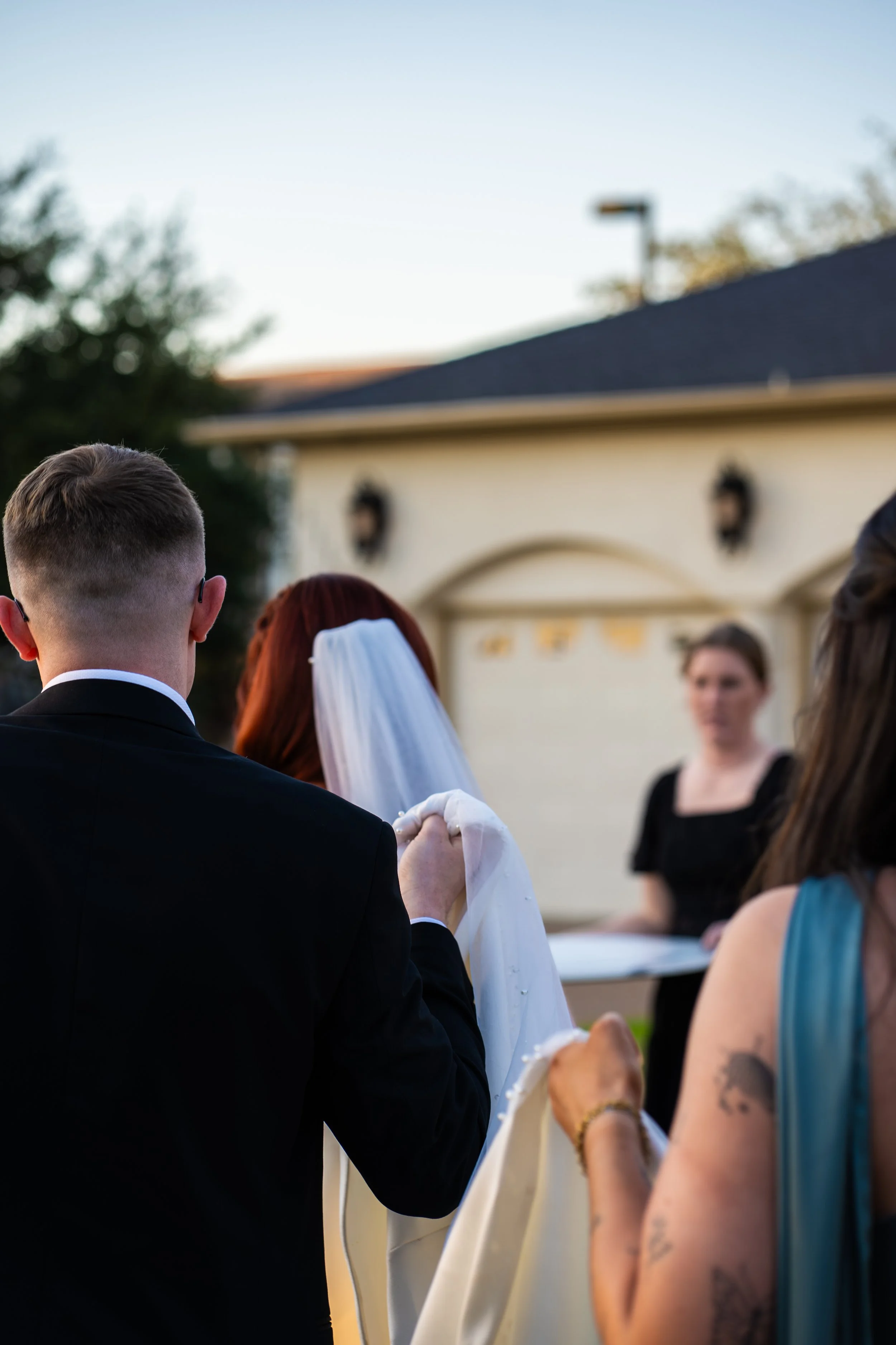 A wedding ceremony taking place outdoors during the daytime with a groom in a black suit and a bride with a veil, holding hands. Several guests are visible, including a woman in a teal dress with arm tattoos, and a woman in black. A house with a gara