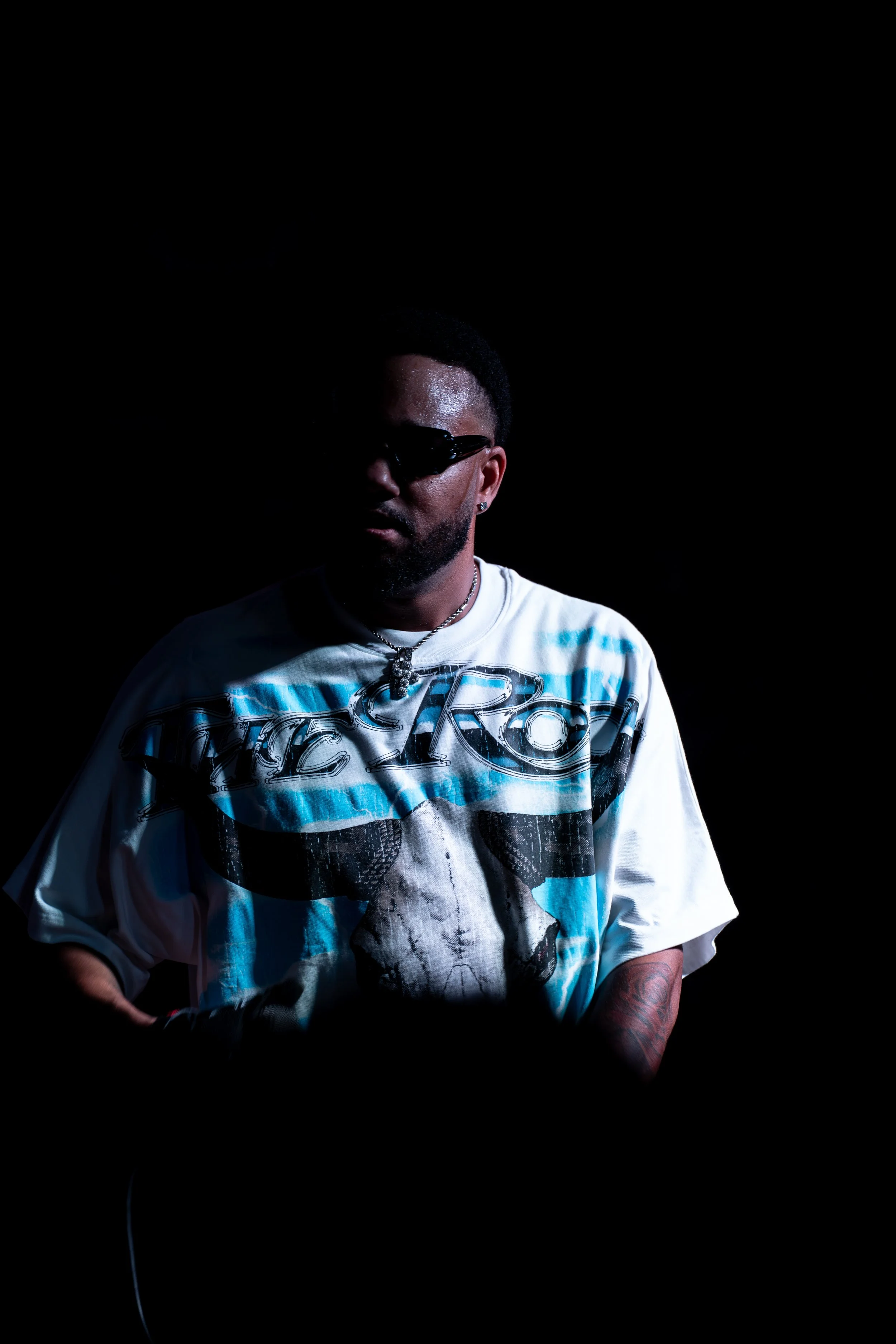 A man with dark skin and a beard wearing sunglasses, a graphic t-shirt, and jewelry facing the camera in a dark setting.