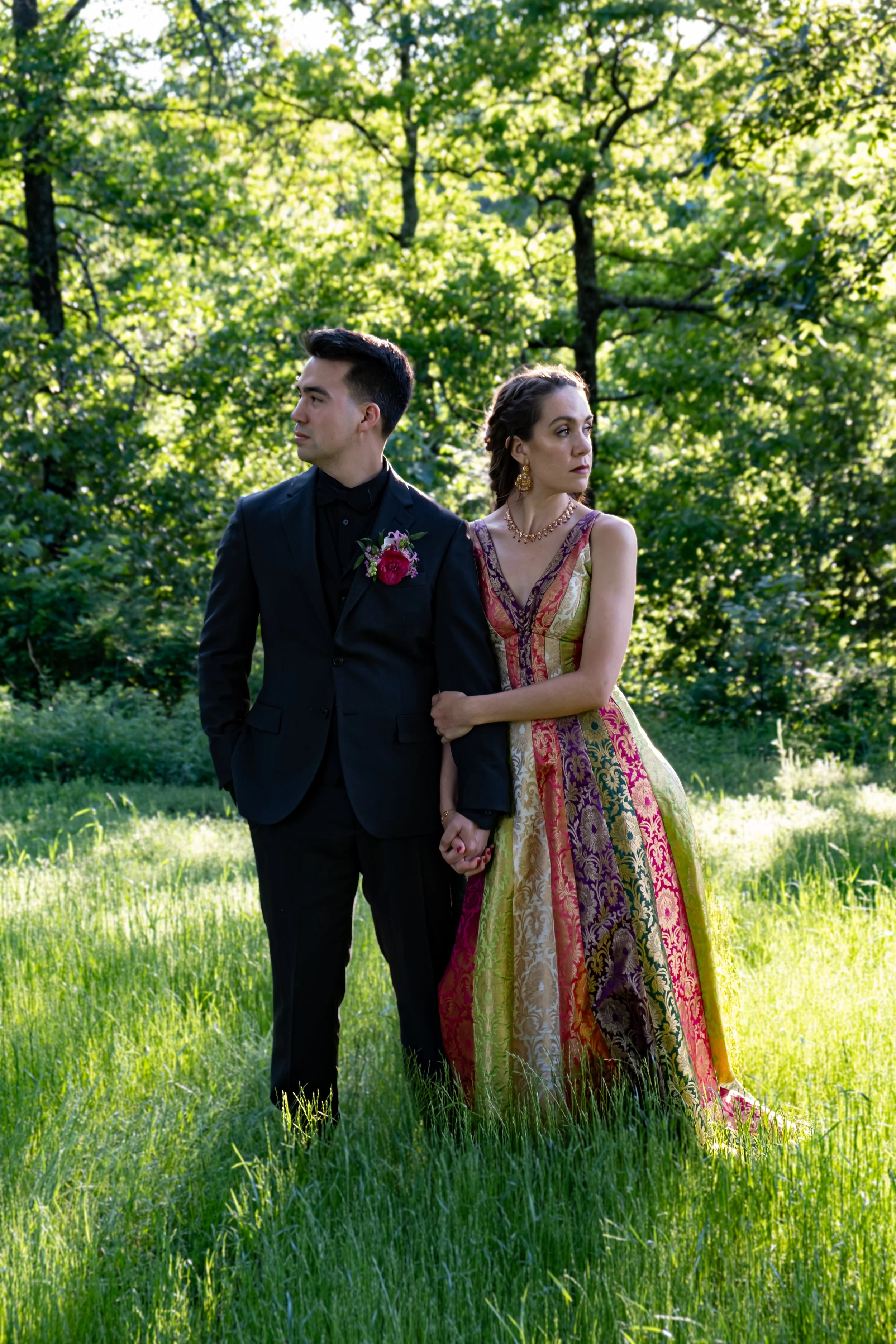 A couple standing outdoors in a grassy area with green trees in the background. The man is wearing a black suit with a boutonniere, and the woman is wearing a colorful, patterned dress with jewelry. They are holding hands and looking in opposite directions.