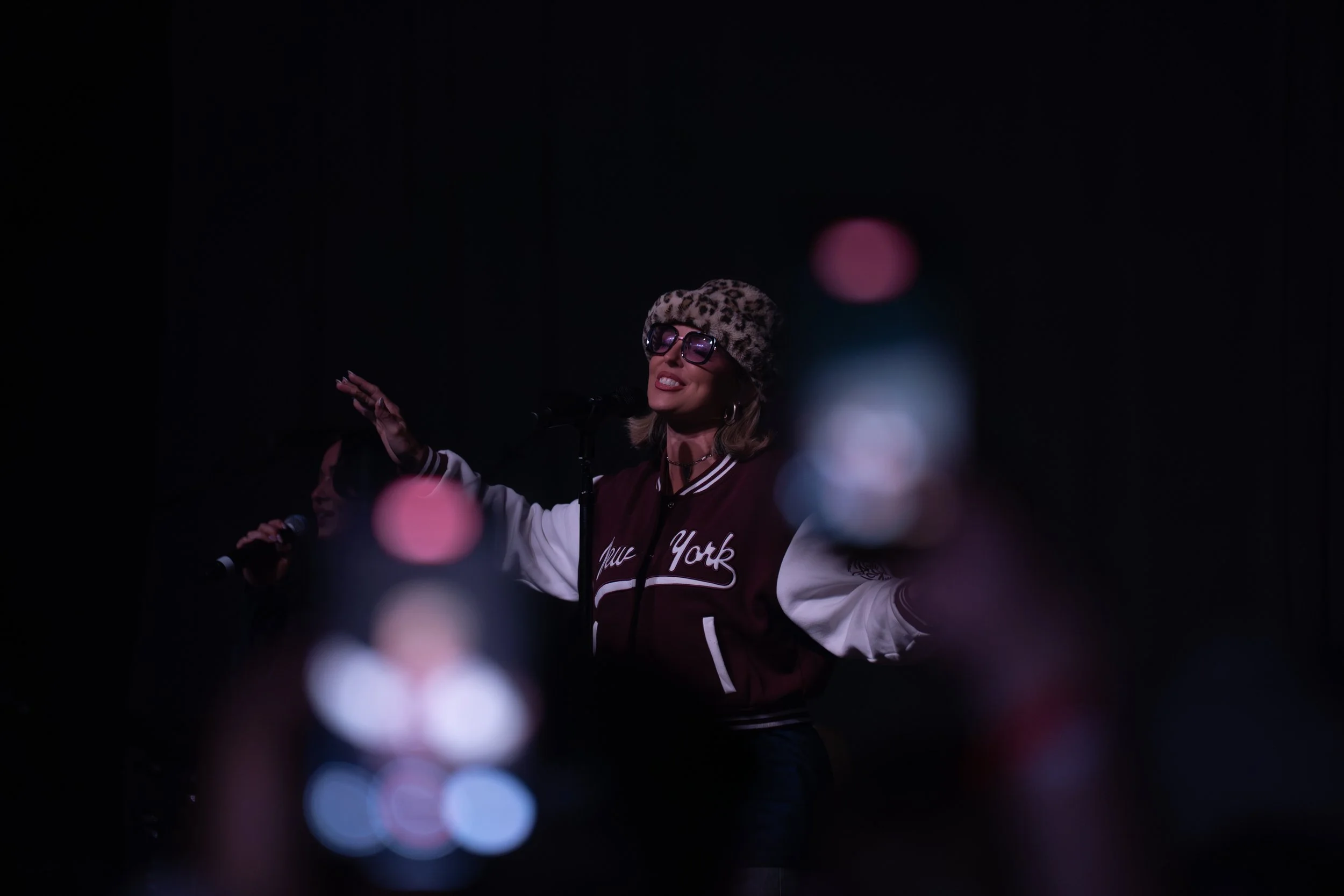 A woman wearing glasses, a leopard print hat, and a jacket with 'New York' written on it, is singing or speaking into a microphone on a dark stage with colorful out-of-focus lights in the foreground.