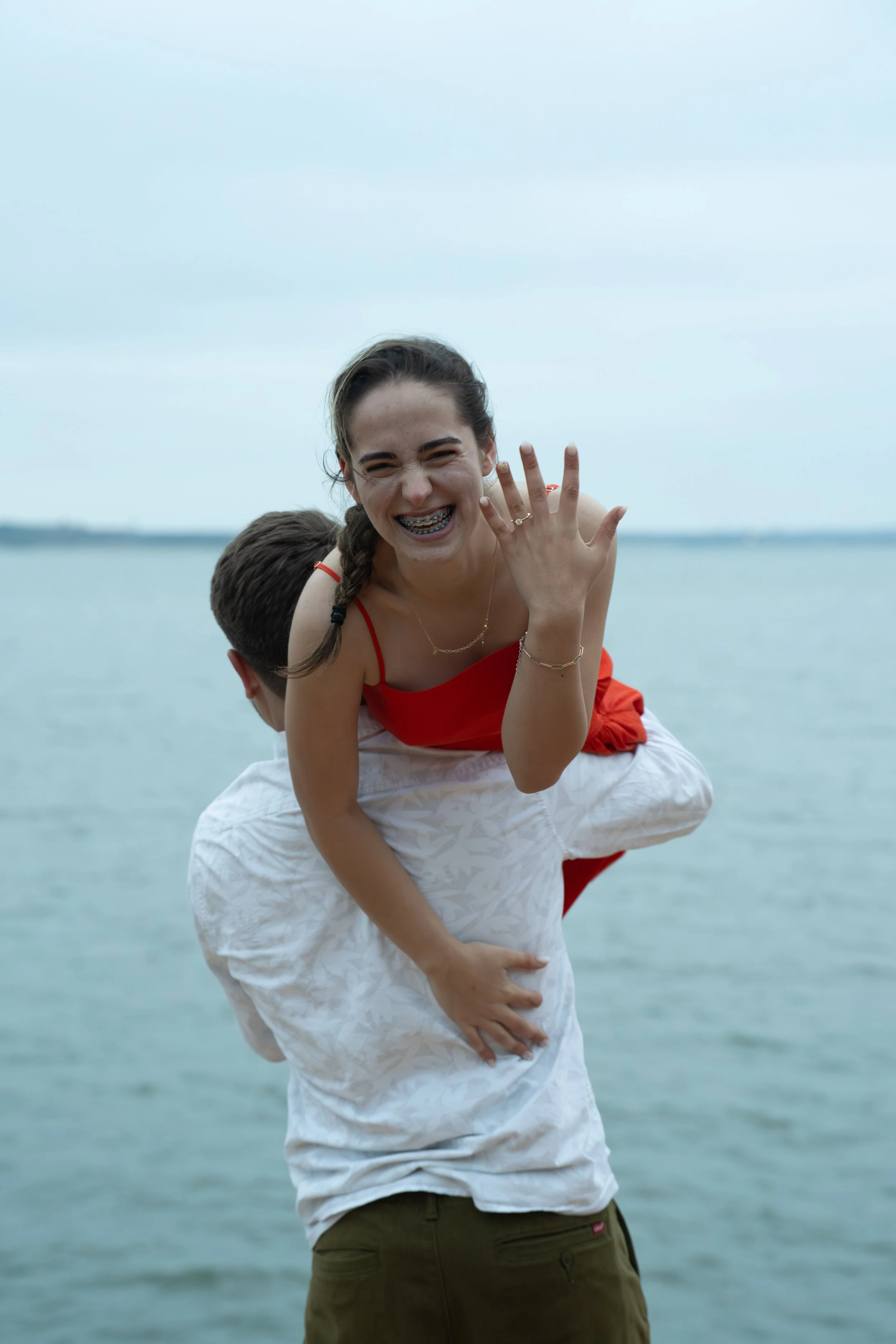 A smiling young woman with braces, wearing a red tank top, and a man with glasses and a white shirt, standing near a body of water on a cloudy day.