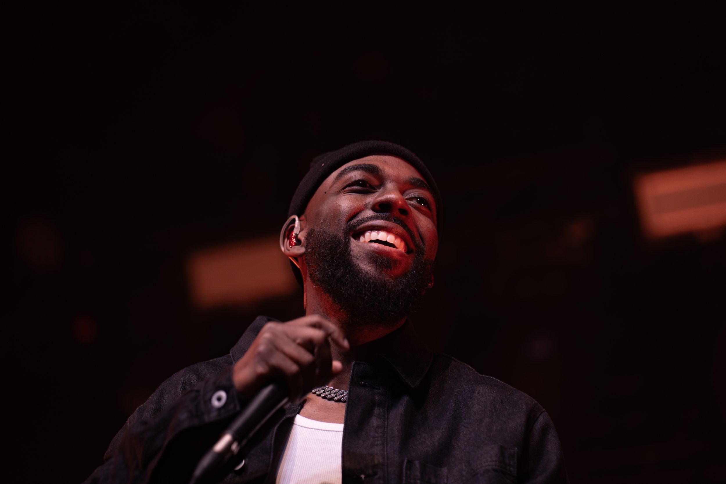 A man smiling and holding a microphone. He is wearing a black jacket, a chain necklace, and a black beanie, with a dark background.