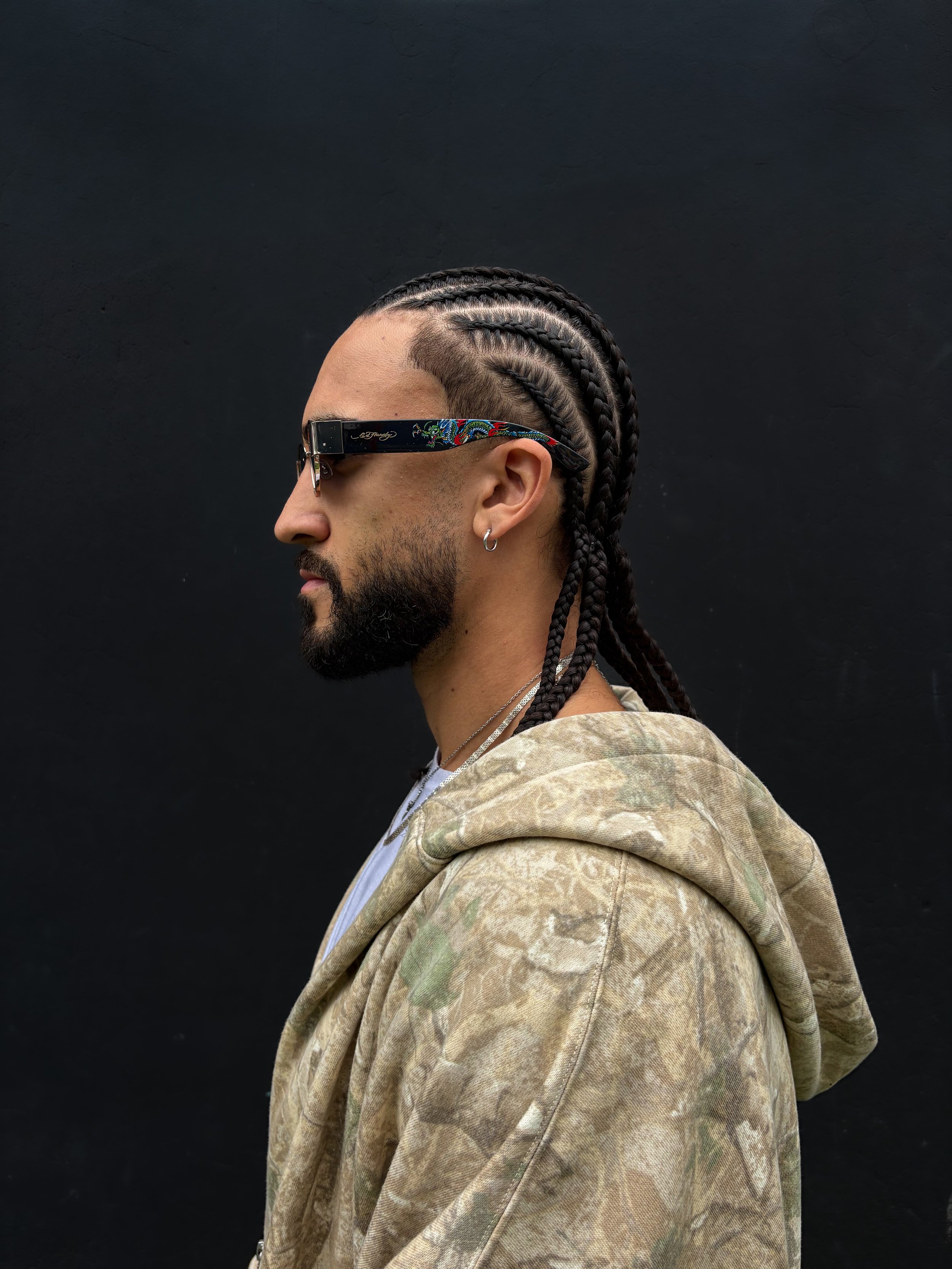 Side profile of a man with braided hair wearing sunglasses, earrings, and a beige camouflage jacket against a black background.