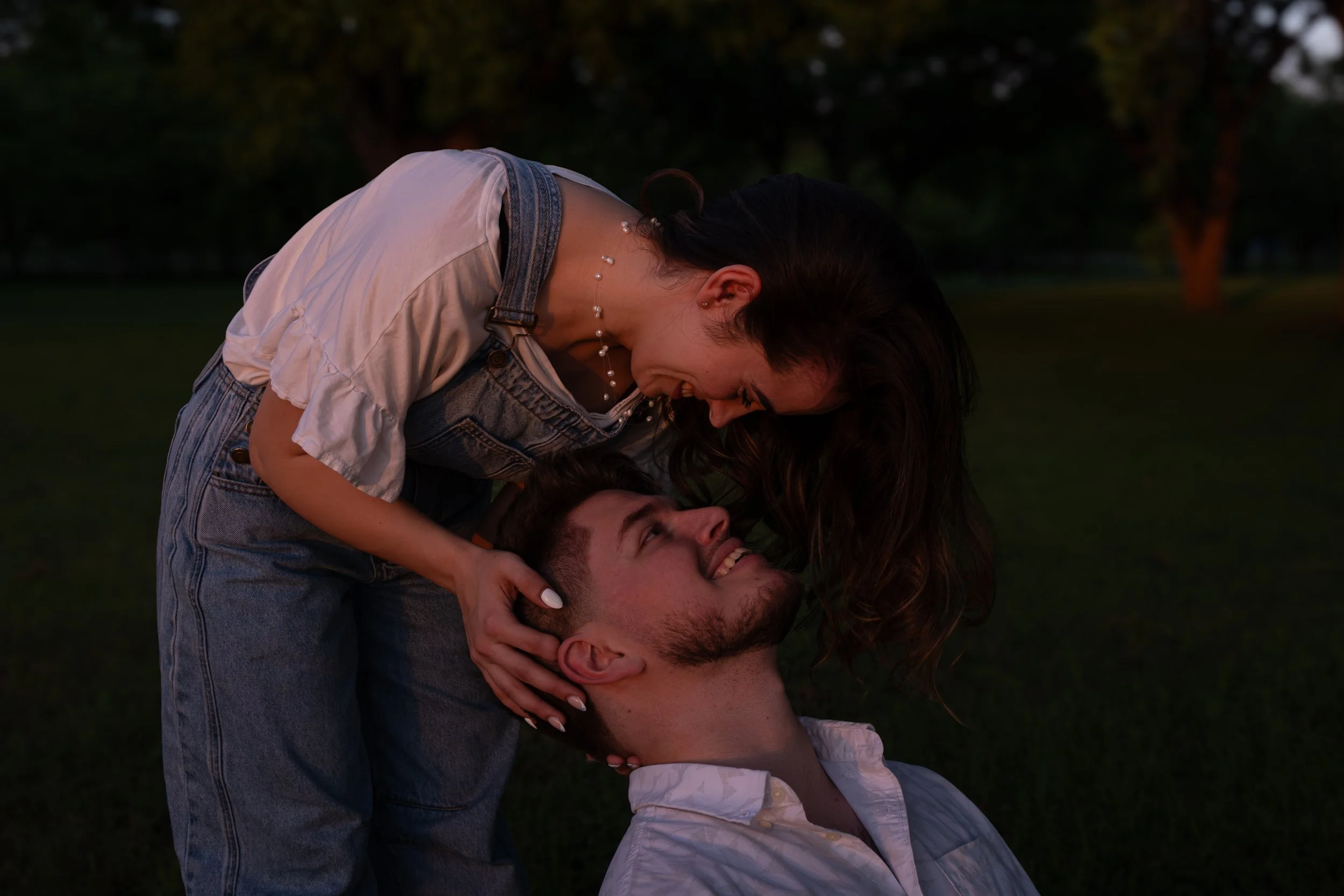 A woman and a man smiling and playing together outdoors in a park at sunset, with trees in the background.
