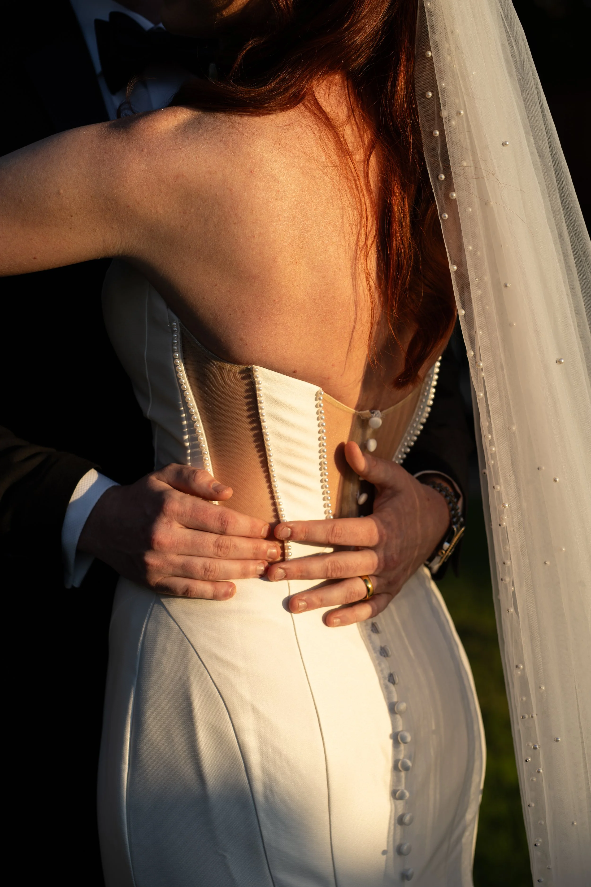 A bride in a white wedding gown with pearl accents, wearing a veil, being embraced from behind by a man in a black tuxedo. The man’s hands are on the bride’s waist and stomach.