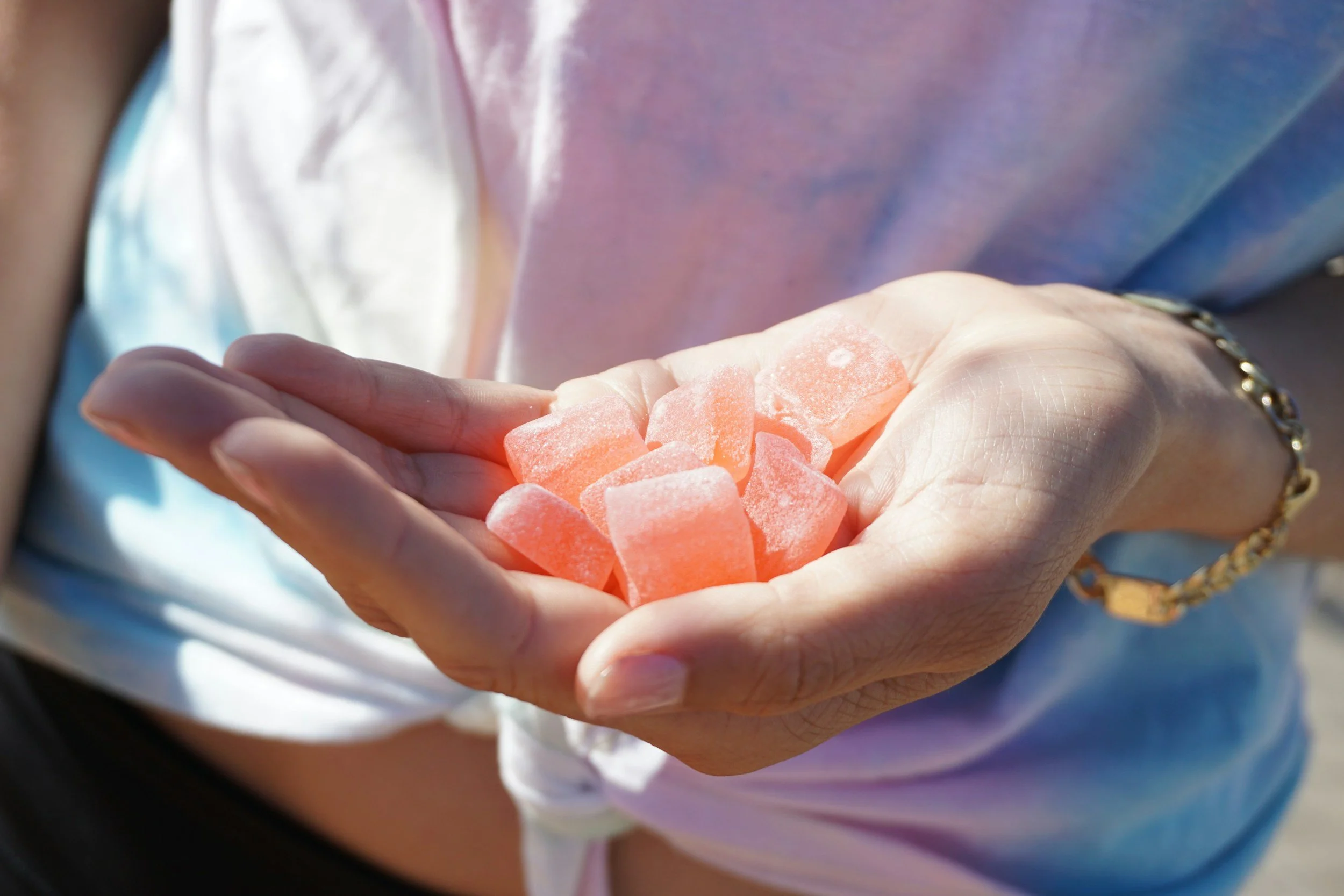 A hand holding pink candy gummy squares outdoors, with sunlight and a person wearing a pastel tie-dye shirt.