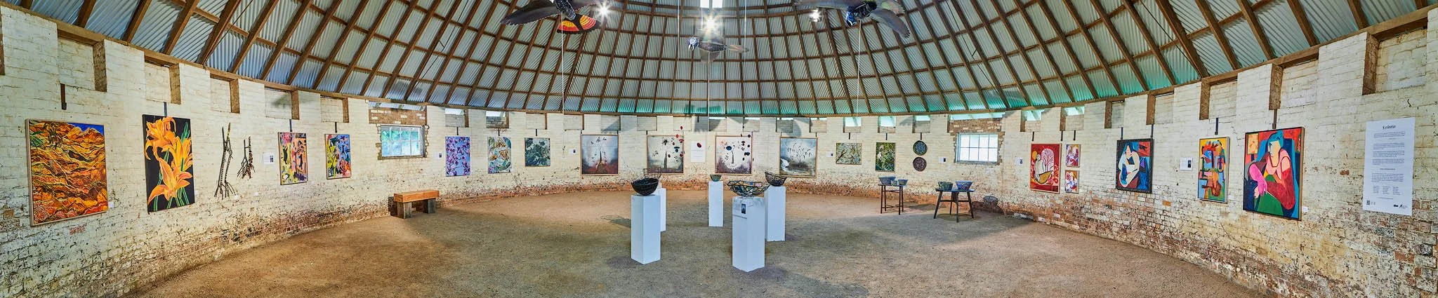An art gallery with colorful paintings on the brick wall and sculptures on white pedestals inside a round building with a wooden ceiling.