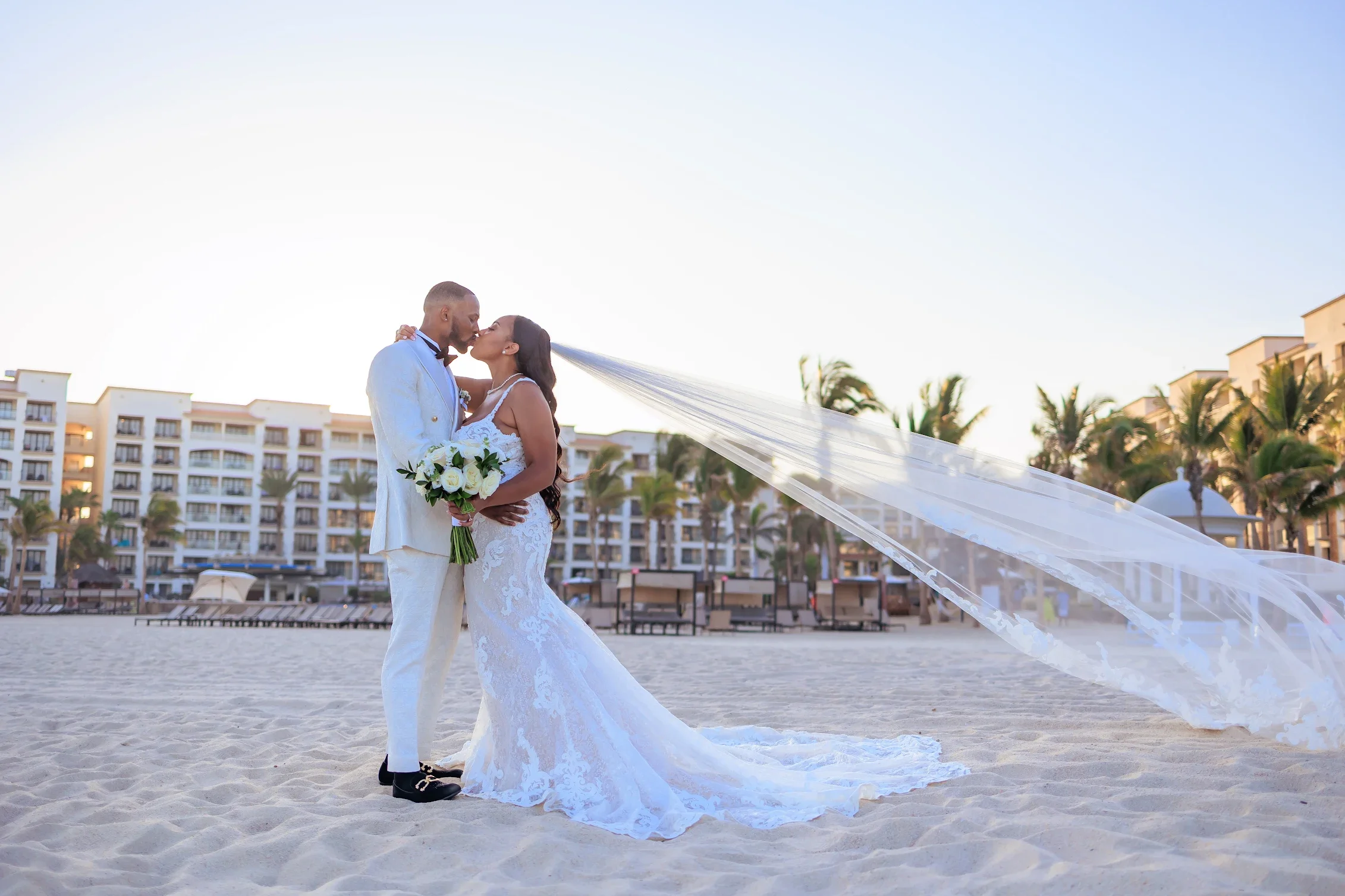 A bride and groom kiss on the beach during their wedding, with the bride holding a bouquet of white roses and a flowing veil.