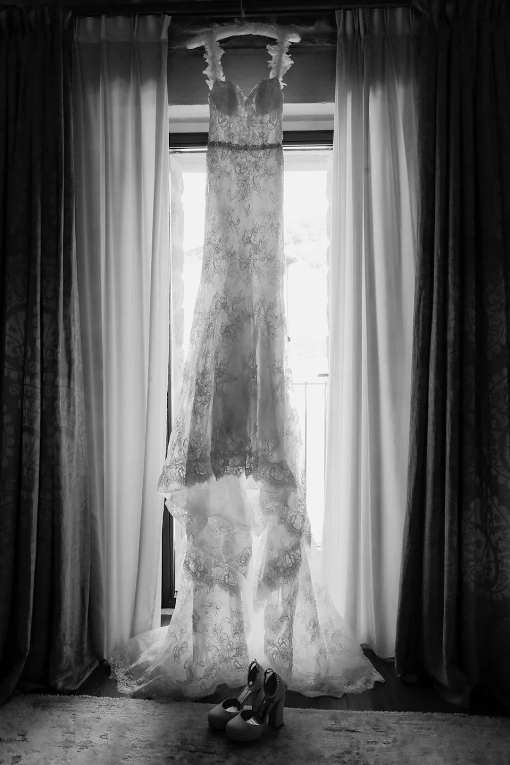 A wedding dress hangs in front of a window with curtains, with a pair of high-heeled shoes placed underneath.