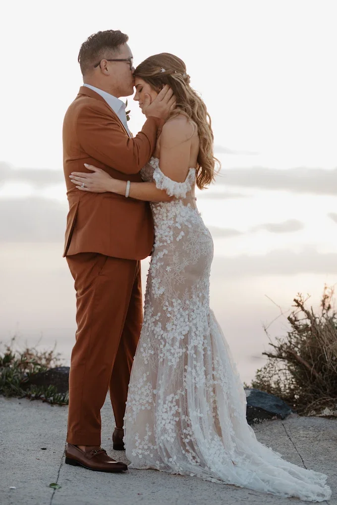 A wedding couple hugging on a beach at sunset, with the man in a brown suit and the woman in a white lace wedding dress.