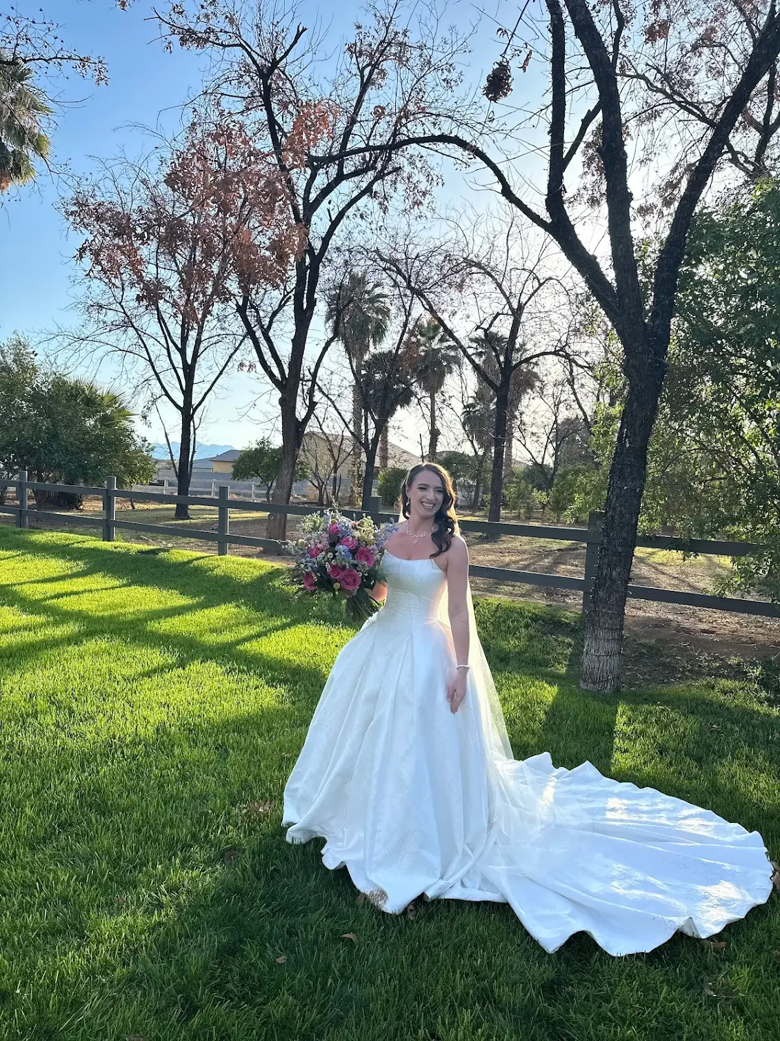 A bride in a white wedding dress holding a bouquet of flowers, standing on green grass with trees and a fence in the background, smiling in sunlight.