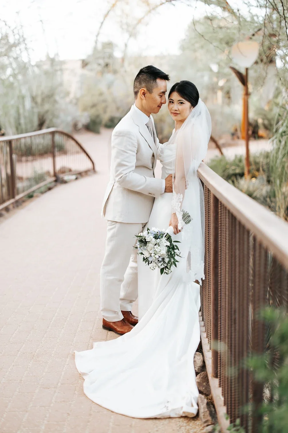 A bride in a white wedding dress and veil holding a bouquet stands next to a groom in a white suit on a bridge outdoors during sunset.