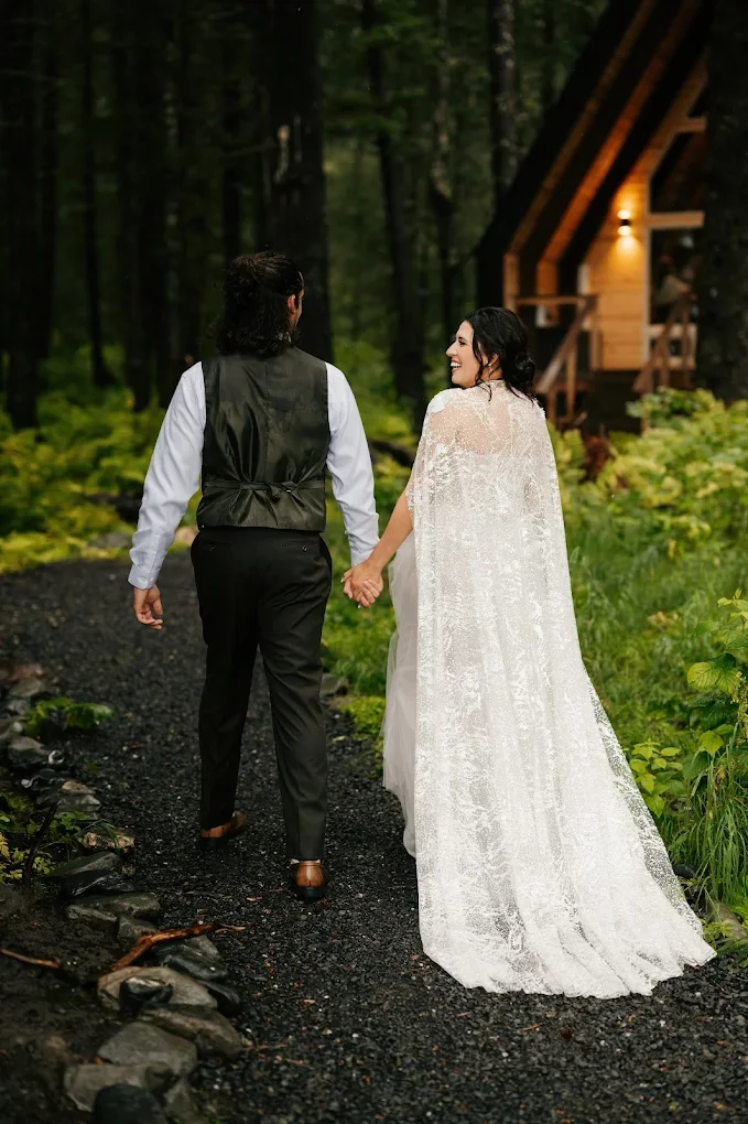 A newlywed couple walking hand in hand through a forest with lush green foliage, a gravel path, and a wooden cabin in the background, smiling at each other.