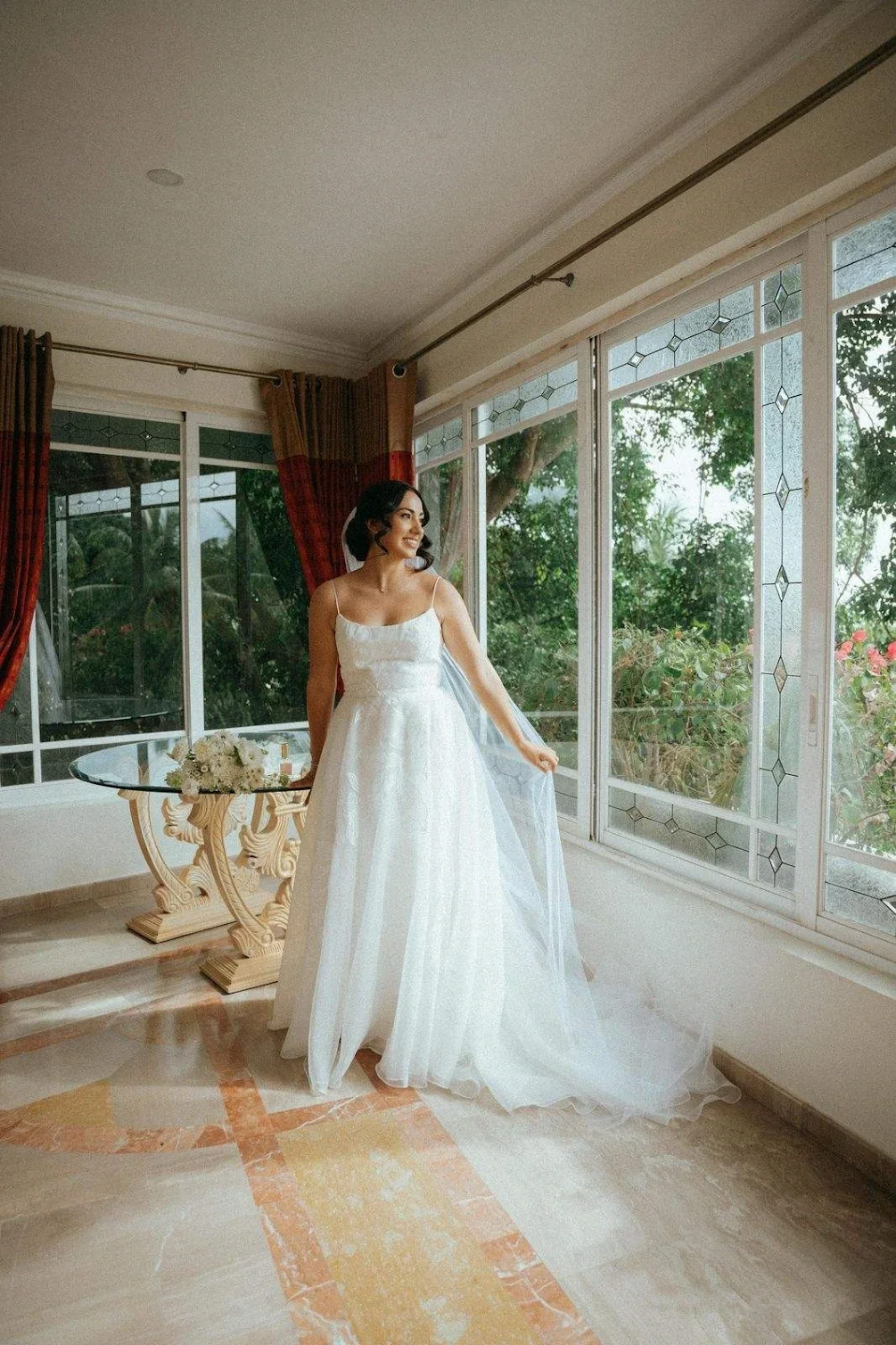 A woman in a white wedding dress standing indoors near large windows with greenery outside, holding her dress and smiling.