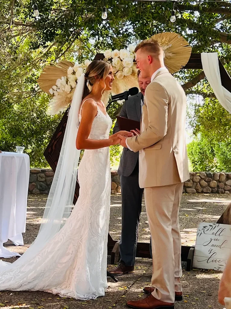 A bride and groom exchanging vows during an outdoor wedding ceremony, with an officiant standing between them, under a decorated canopy of flowers and greenery.
