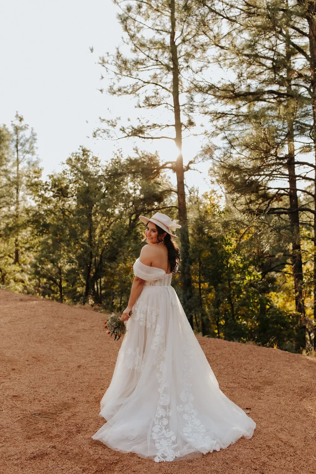 A woman in a white off-shoulder wedding dress and a wide-brimmed hat holding a small bouquet, standing on a dirt path surrounded by tall trees in a forest during sunset.