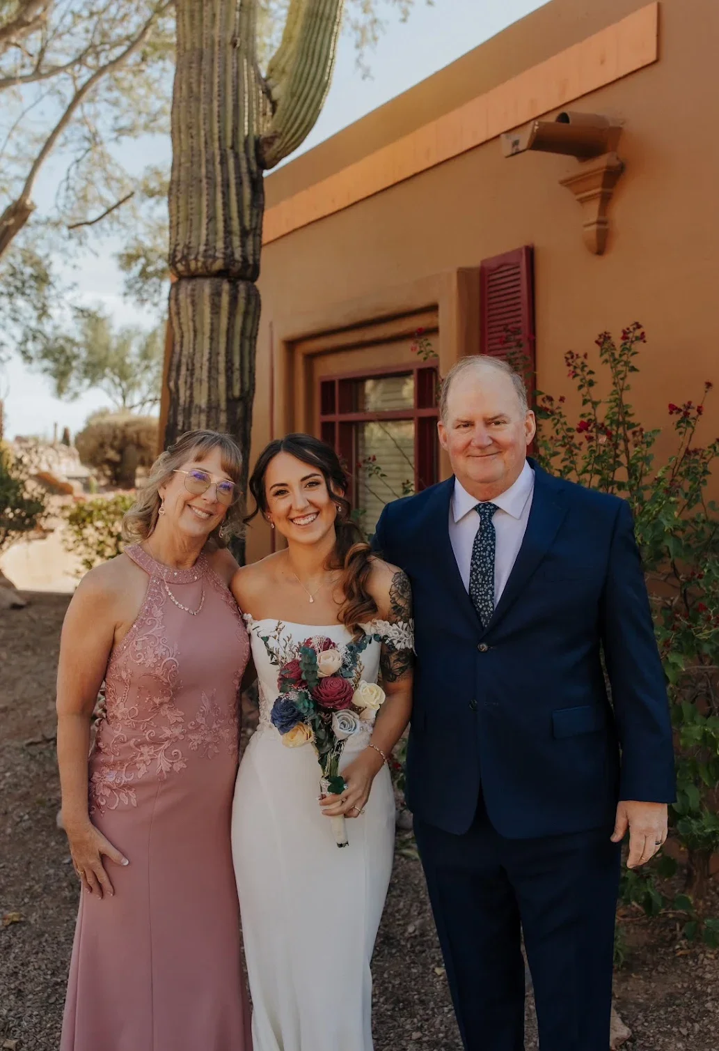 Three people, a woman in a pink gown, a bride in a white wedding dress holding a bouquet, and a man in a navy suit, standing outdoors near a building with a large cactus and desert plants in the background.