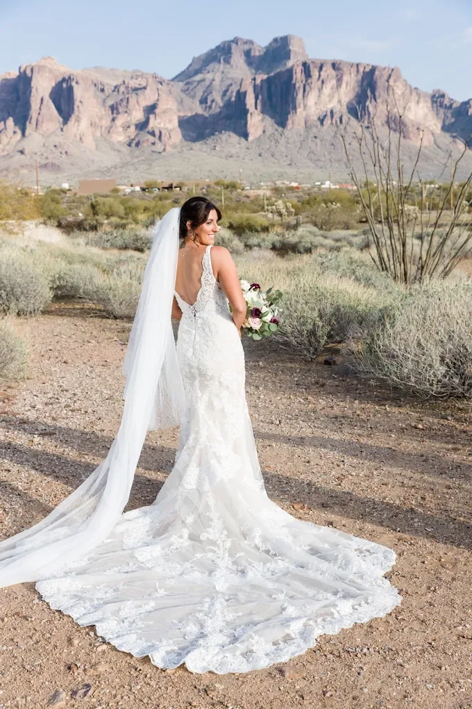 Bride in a white lace wedding dress standing outdoors in a desert landscape with mountains in the background, holding a bouquet of flowers.