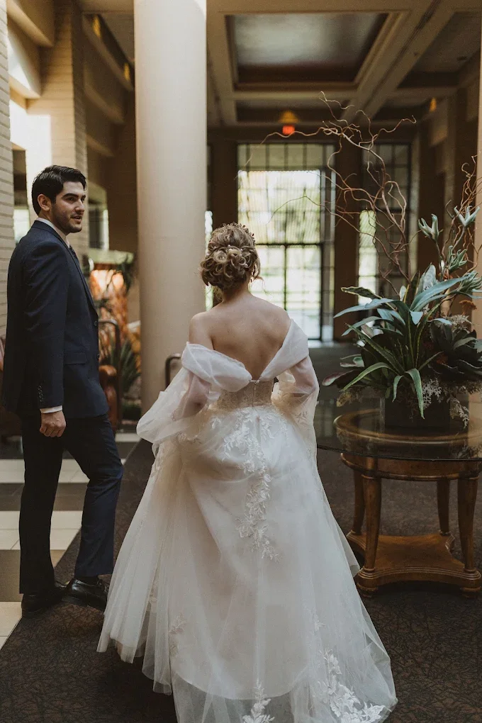Bride in a white wedding gown with off-shoulder sleeves and an updo hairstyle, standing in an elegant lobby area with a man in a dark suit looking at her, next to a large floral arrangement on a wooden table.