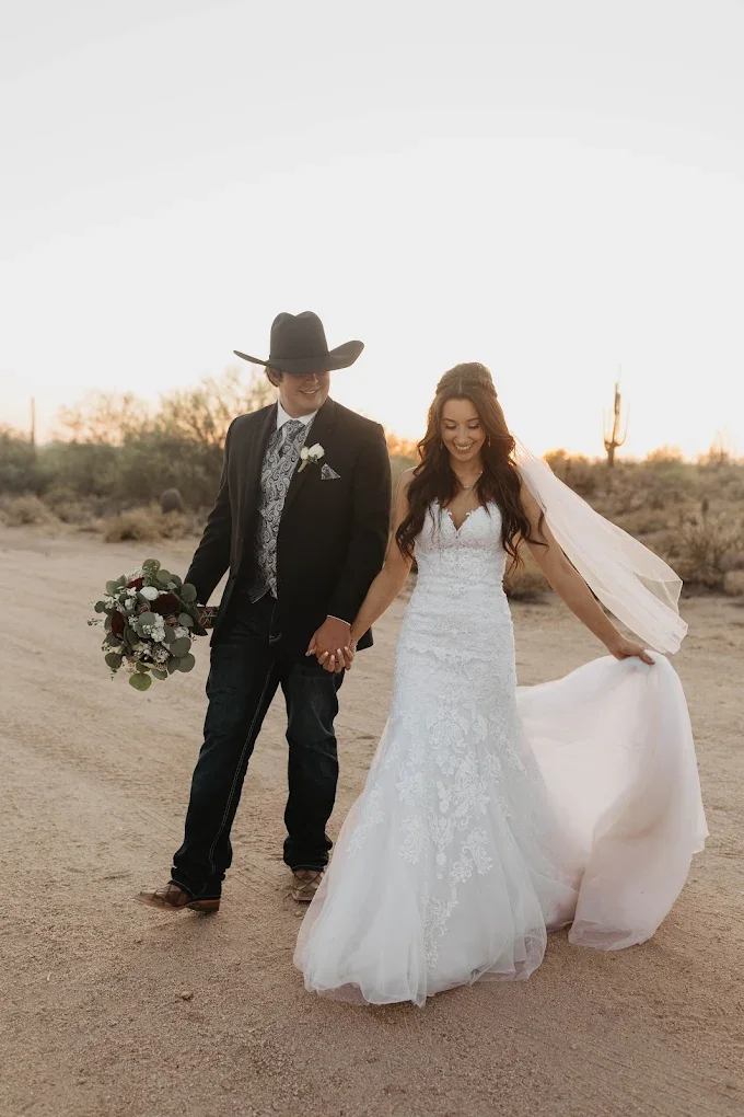 A bride and groom walking hand in hand on desert terrain at sunset, both smiling. The bride wears a white lace wedding gown and a veil, holding the train of her dress, while the groom wears a dark suit, a cowboy hat, and holds a bouquet of flowers.