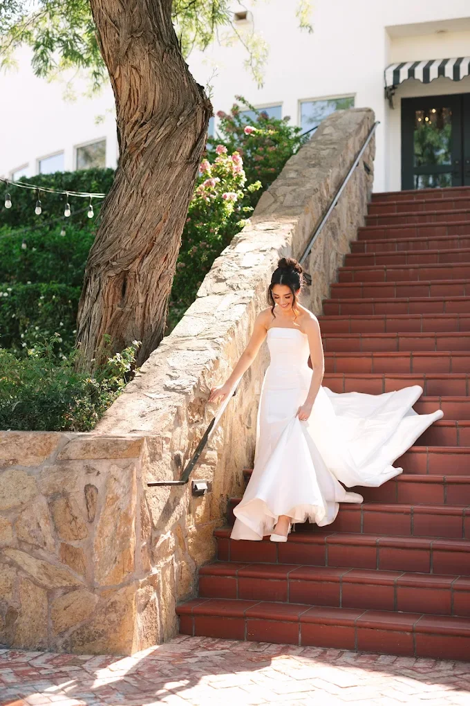 A bride in a white wedding dress walking down outdoor stairs with a stone wall, a large tree, and greenery.