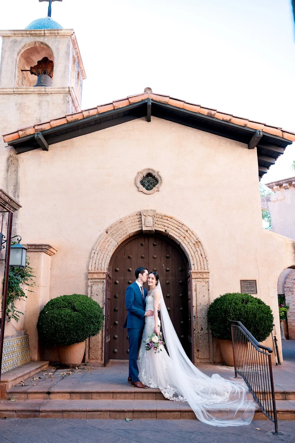 A bride and groom in wedding attire standing in front of a rustic building with a large wooden door and a bell tower with a dome in the background.