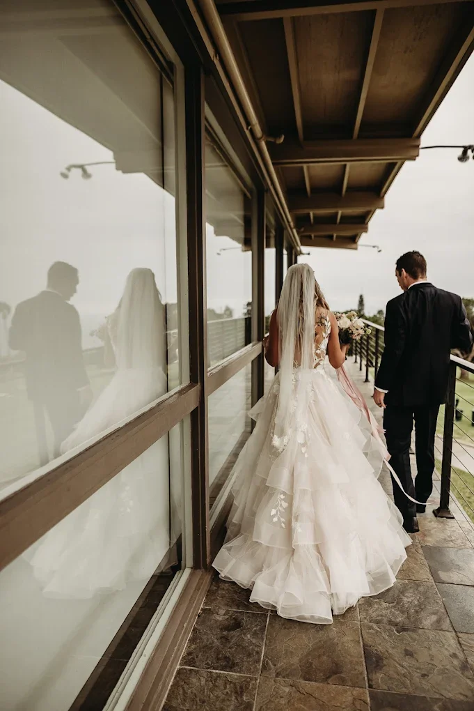 Bride in a white wedding gown with a long veil walking on a balcony accompanied by a groom in a black suit