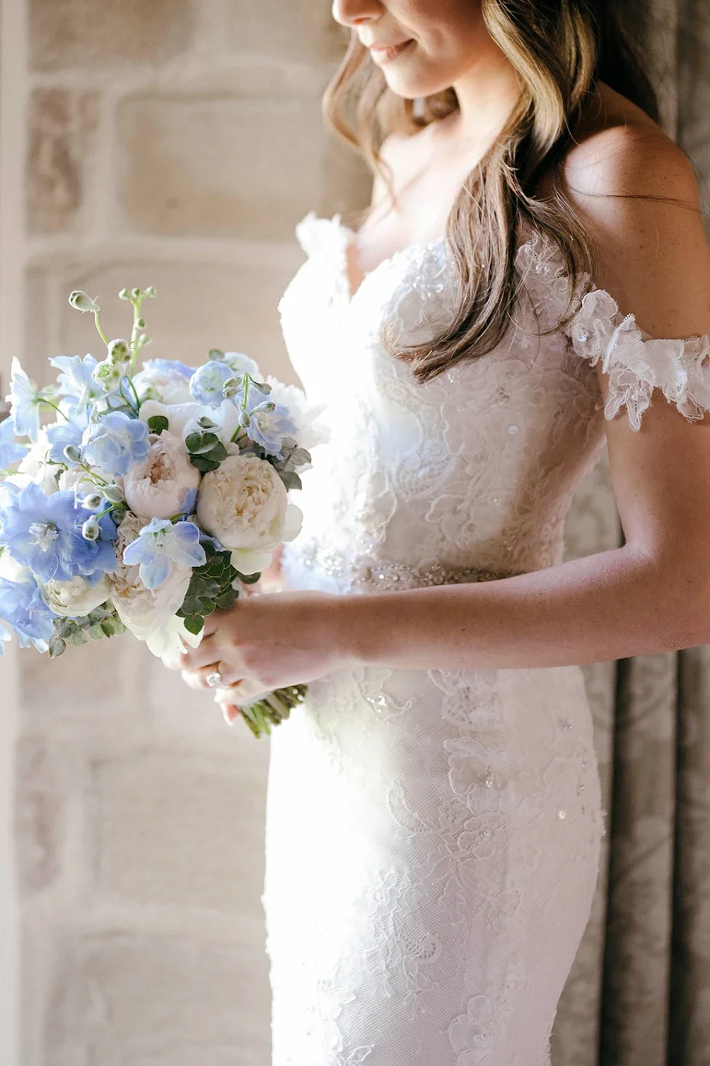 Close-up of a bride in a lace wedding gown holding a bouquet of white and light blue flowers, with a brick wall in the background.