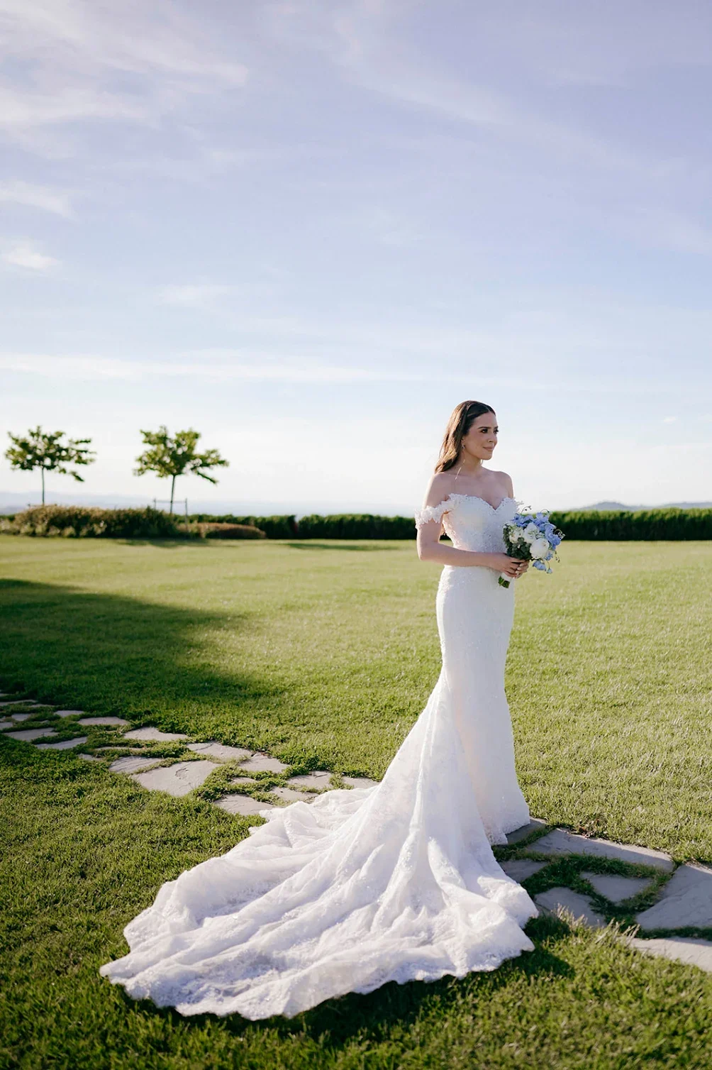 Bride in a white lace wedding gown holding a bouquet of flowers outdoors in a grassy field with a clear blue sky.