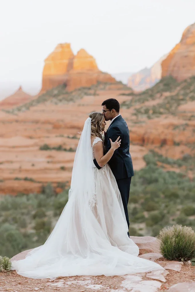 A newlywed couple stands close together on a rock formation, with red rock mountains in the background. The bride wears a white wedding dress with a long train and veil, and the groom wears a dark suit. They appear to be sharing a kiss in a desert landscape.