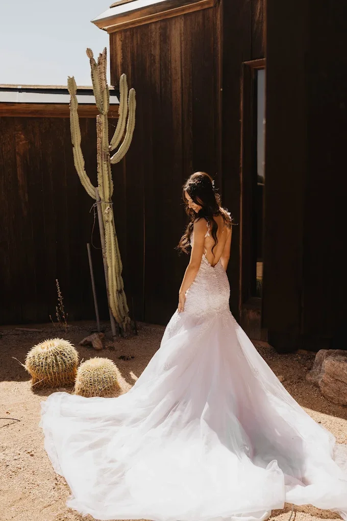 Brunette woman in a white lace wedding dress standing outdoors in a desert setting with cacti, against a dark wooden wall.