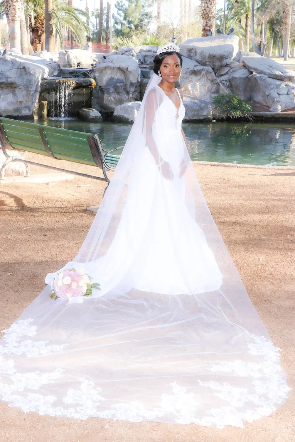 A woman in a wedding dress with a long train and veil, standing near a pond with rocks and waterfall, holding a bouquet of flowers.