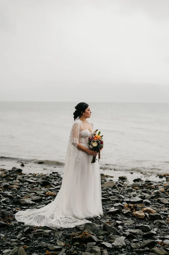 Brunette woman in a white wedding dress holding a bouquet of flowers standing on a rocky beach near the ocean, facing sideways.