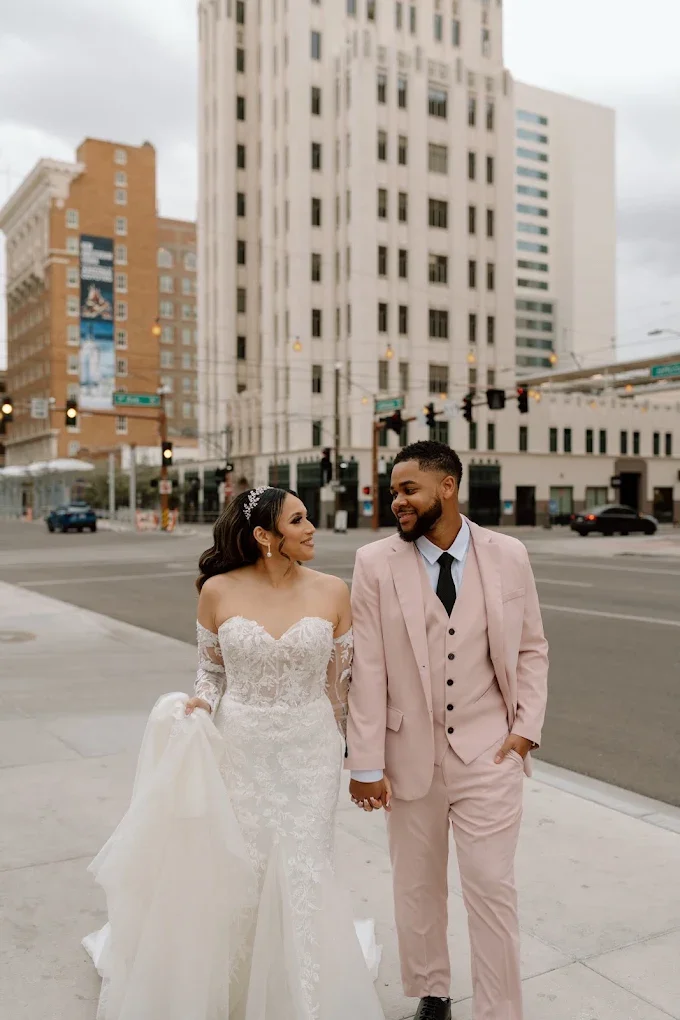 Bride and groom walking hand in hand on city sidewalk, smiling at each other, with tall buildings and traffic lights in the background.