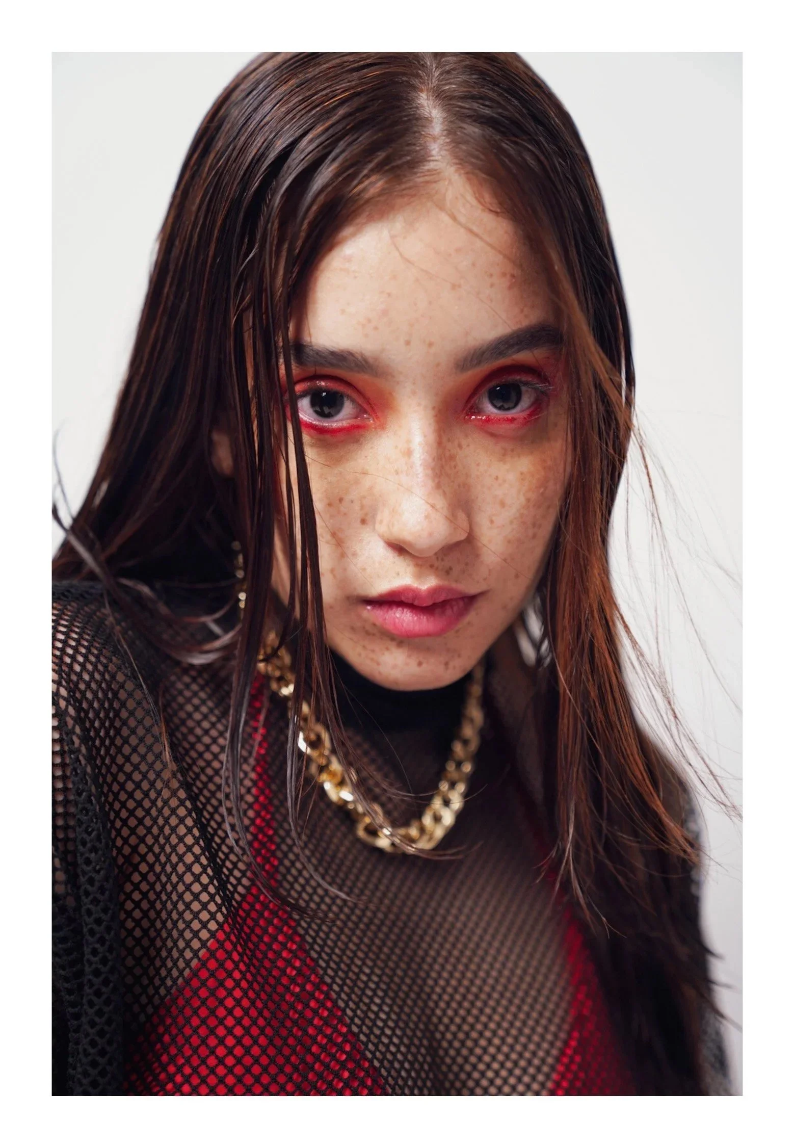 Close-up of a young woman with wet, reddish-brown hair, red eye makeup, freckles, and a serious expression, wearing a black mesh top, red garment underneath, and a chunky gold chain necklace.