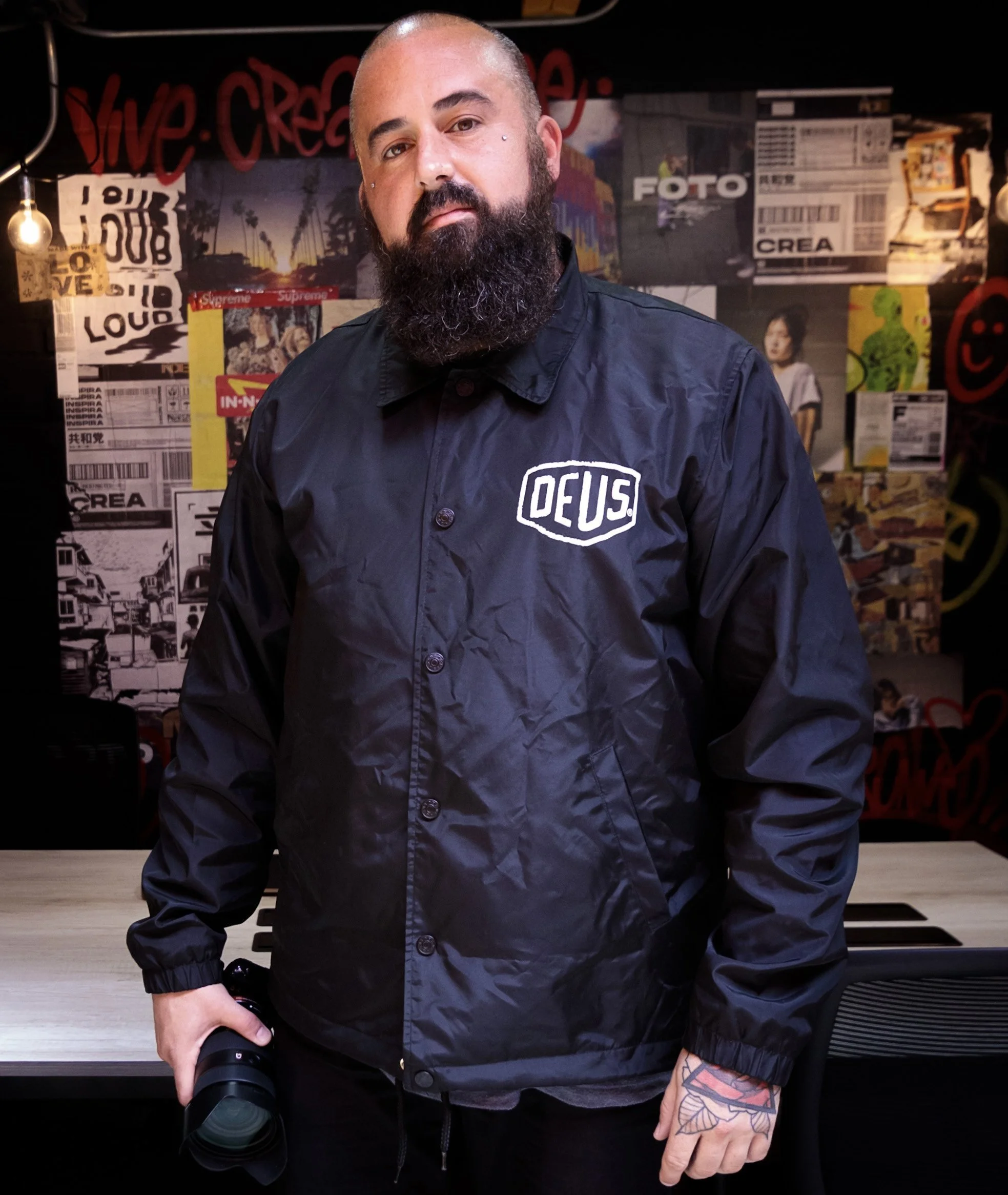 Man with a beard, holding a camera, standing in front of a collage wall with posters and graffiti, wearing a dark jacket with a white "DEUS" patch.