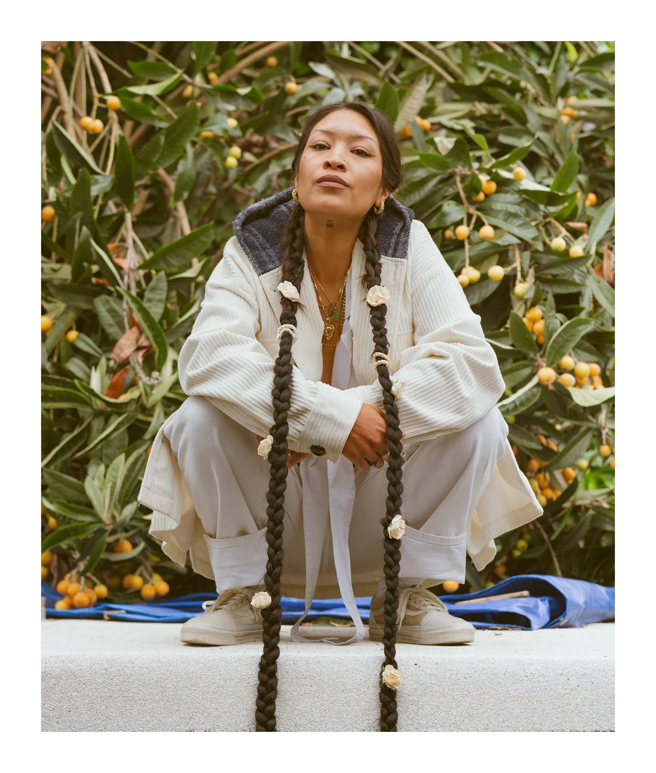 Woman with long rope braids crouching on a white ledge in a cream jacket in front of a loquat tree heavy with fruit.