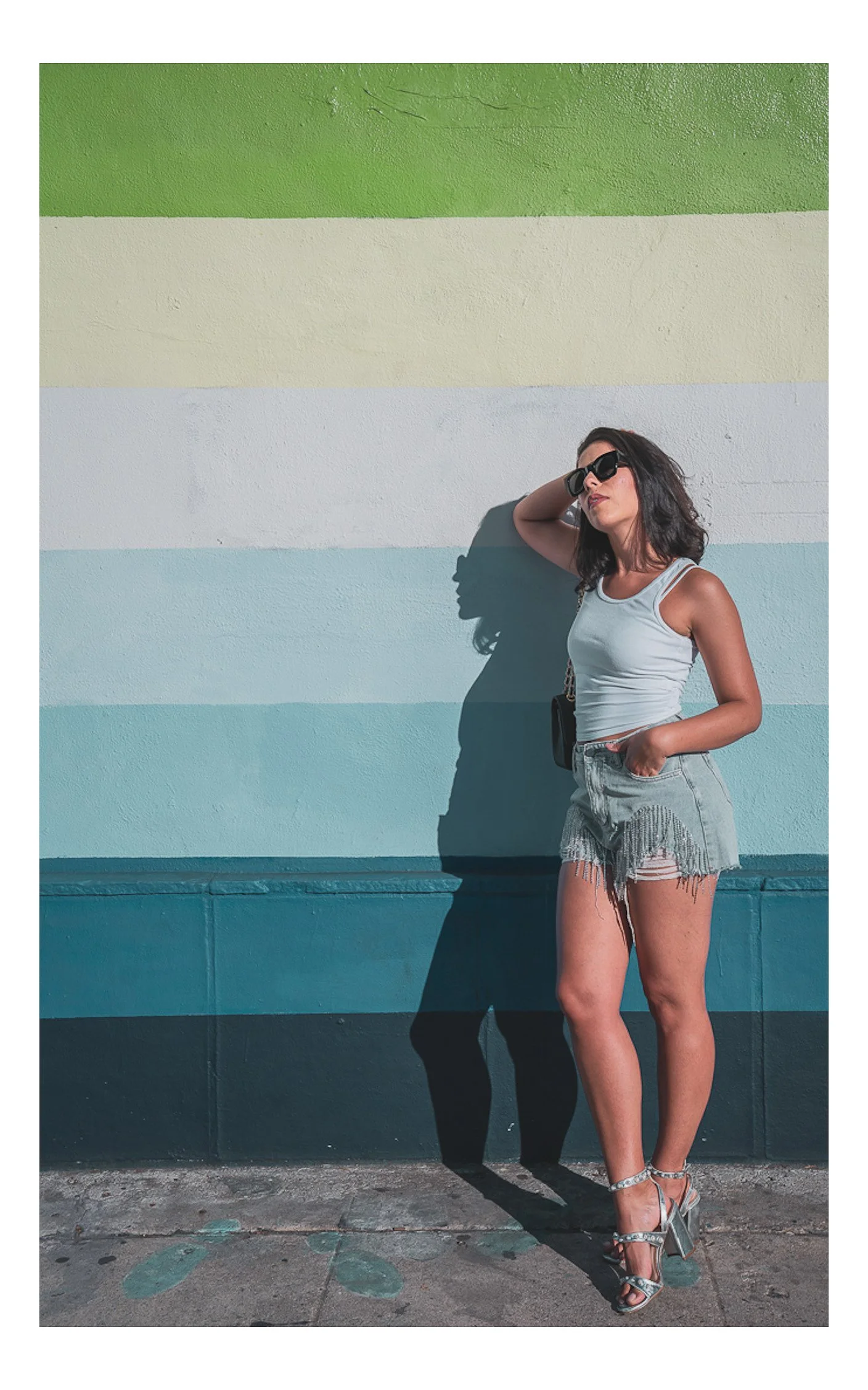 Woman in sunglasses, white tank top and fringed shorts against a striped teal wall