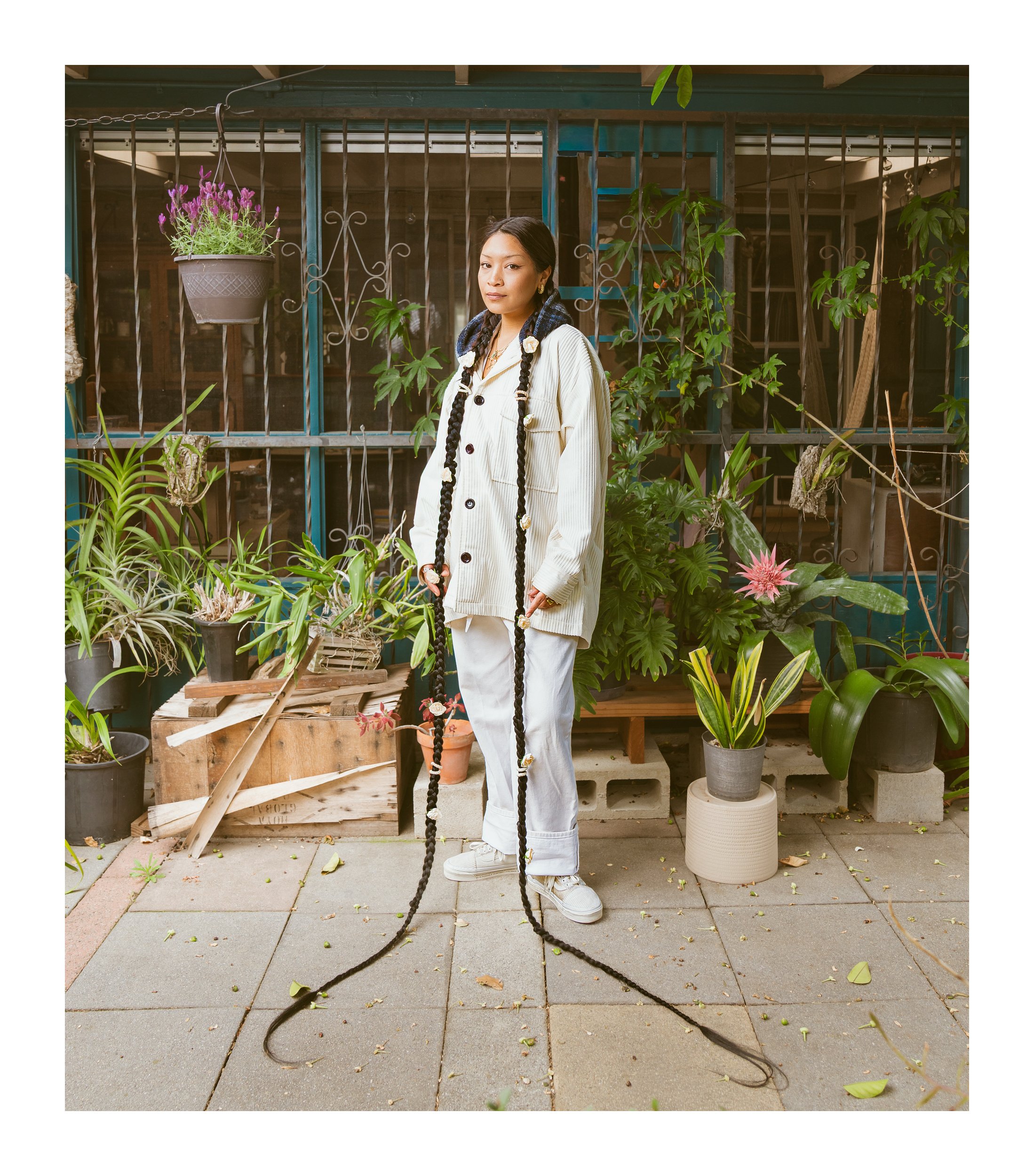 Young woman with long black braids in a white jacket and pants standing among potted plants on a patio.