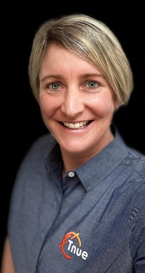 A woman with short blonde hair, wearing a blue collared shirt with an embroidered logo that says "True" and a stylized orange and yellow arrow.