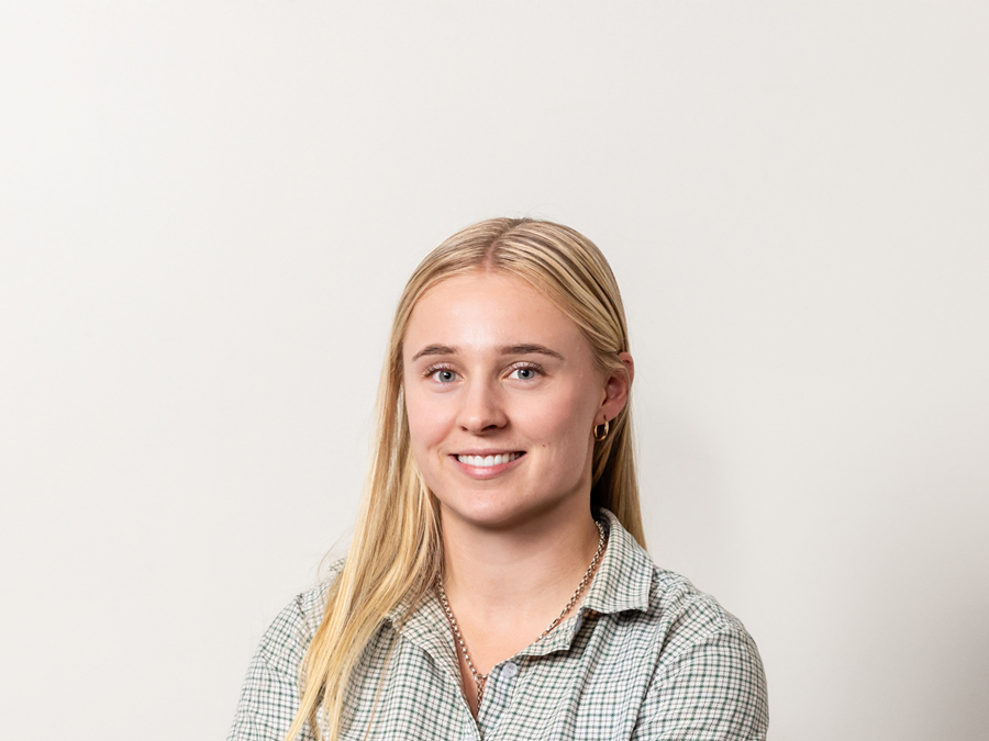 Portrait of a young woman with long blond hair, wearing a checkered shirt and gold jewelry, smiling at the camera against a plain white background.