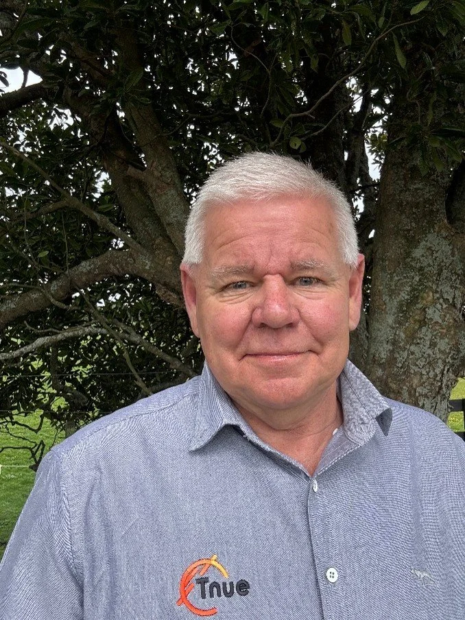 A man with short gray hair and a light gray button-up shirt standing outdoors in front of a tree with green leaves.