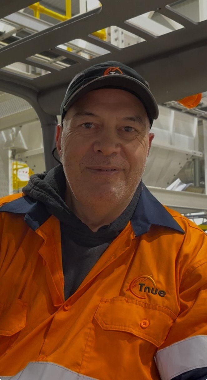 A man in an industrial uniform wearing an orange safety jacket and a black cap, inside a factory or industrial setting.