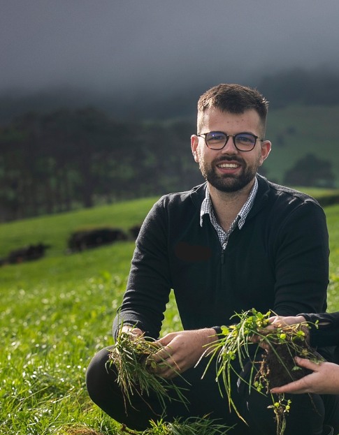 A man with glasses and a beard smiling outdoors in a green field, holding a plant, with hills and cattle in the background.