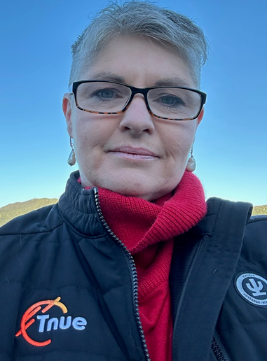 A woman with short gray hair and glasses wearing a red scarf, black jacket with logos, outdoors with a clear blue sky.