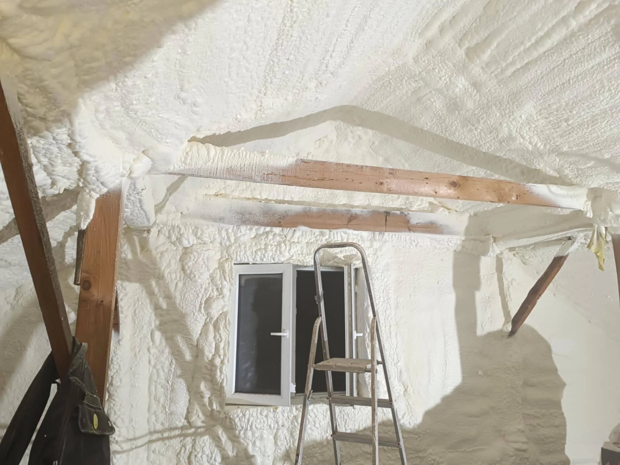 Interior of a room under construction with foam insulation on walls, a ladder, window, and wooden beams.