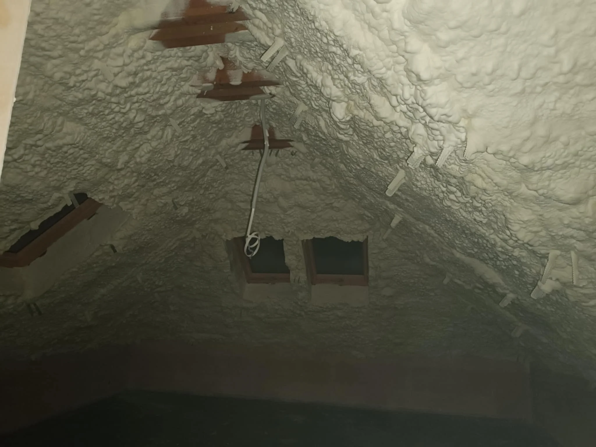 Attic ceiling with spray foam insulation and three small windows, some wooden beams, and an electrical wire hanging down.
